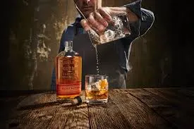 Bulleit Frontier Whiskey being poured from bottle into a glass with ice, with another bottle visible on wooden surface