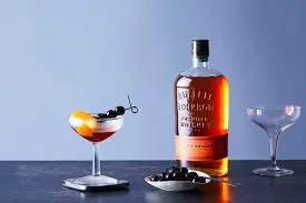 Finished Bulleit Bourbon Manhattan cocktail in a coupe glass, garnished with an orange twist and dark berry, with Bulleit Bourbon bottle visible in the background
